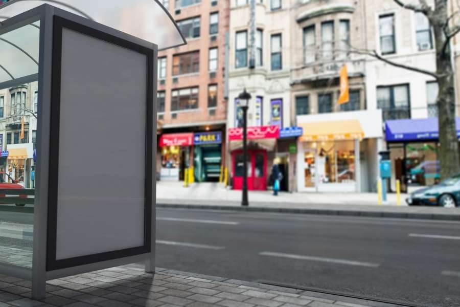 Un panneau publicitaire vertical et vierge installé sous un abribus dans une rue urbaine animée.