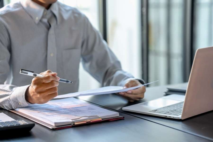 Un homme en chemise tient un stylo et examine des rapports papiers à son bureau, illustrant la gestion des documents administratifs.