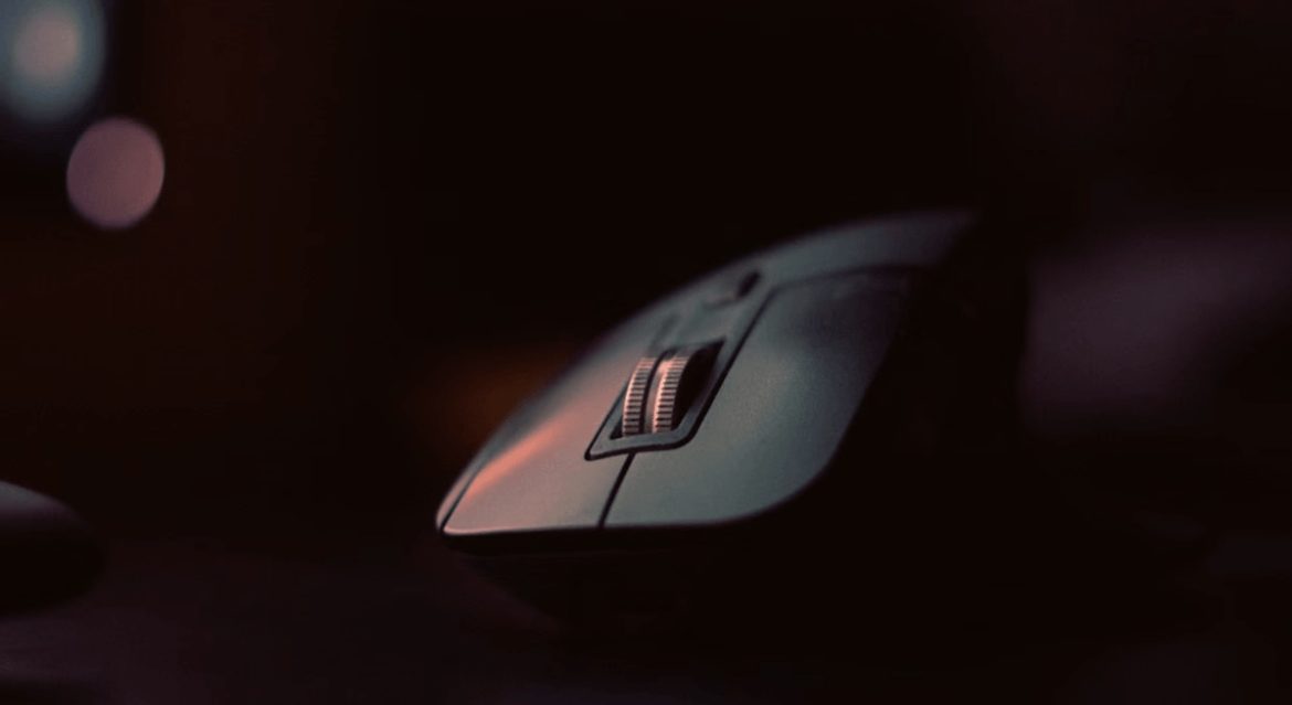 Gros plan d'une souris gaming ergonomique grise avec une molette de défilement métallique en gros plan sur un bureau sombre avec un fond flou.